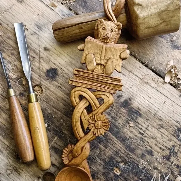 A carving of a custom love spoon depicting a teddy bear sat on books, daisies, and celtic knotwork.